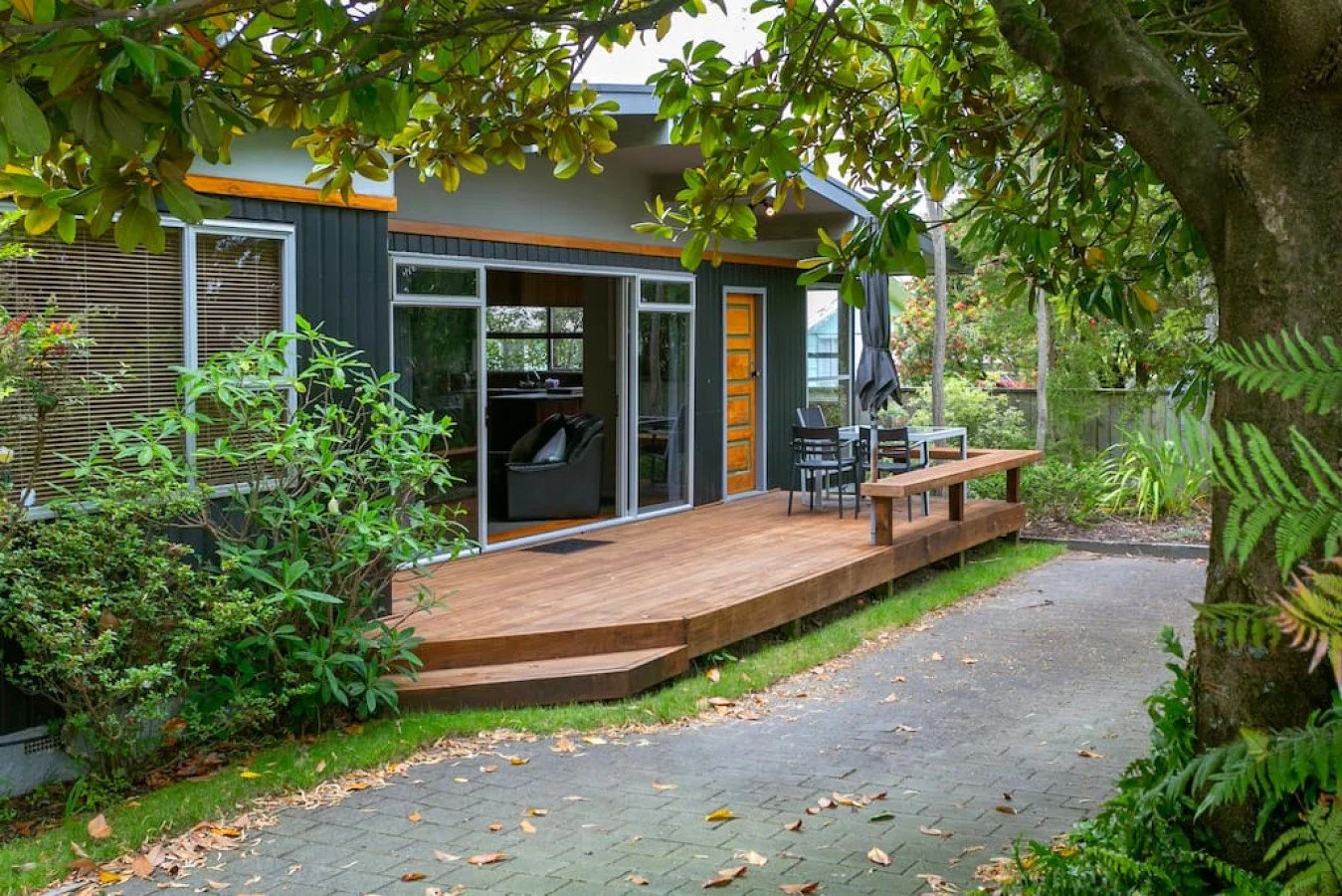 A wooden deck extends from a dark-colored house surrounded by greenery and trees. The deck has outdoor seating with a table and chairs, and is adjacent to a paved pathway. The house features large windows with blinds and a sliding glass door.