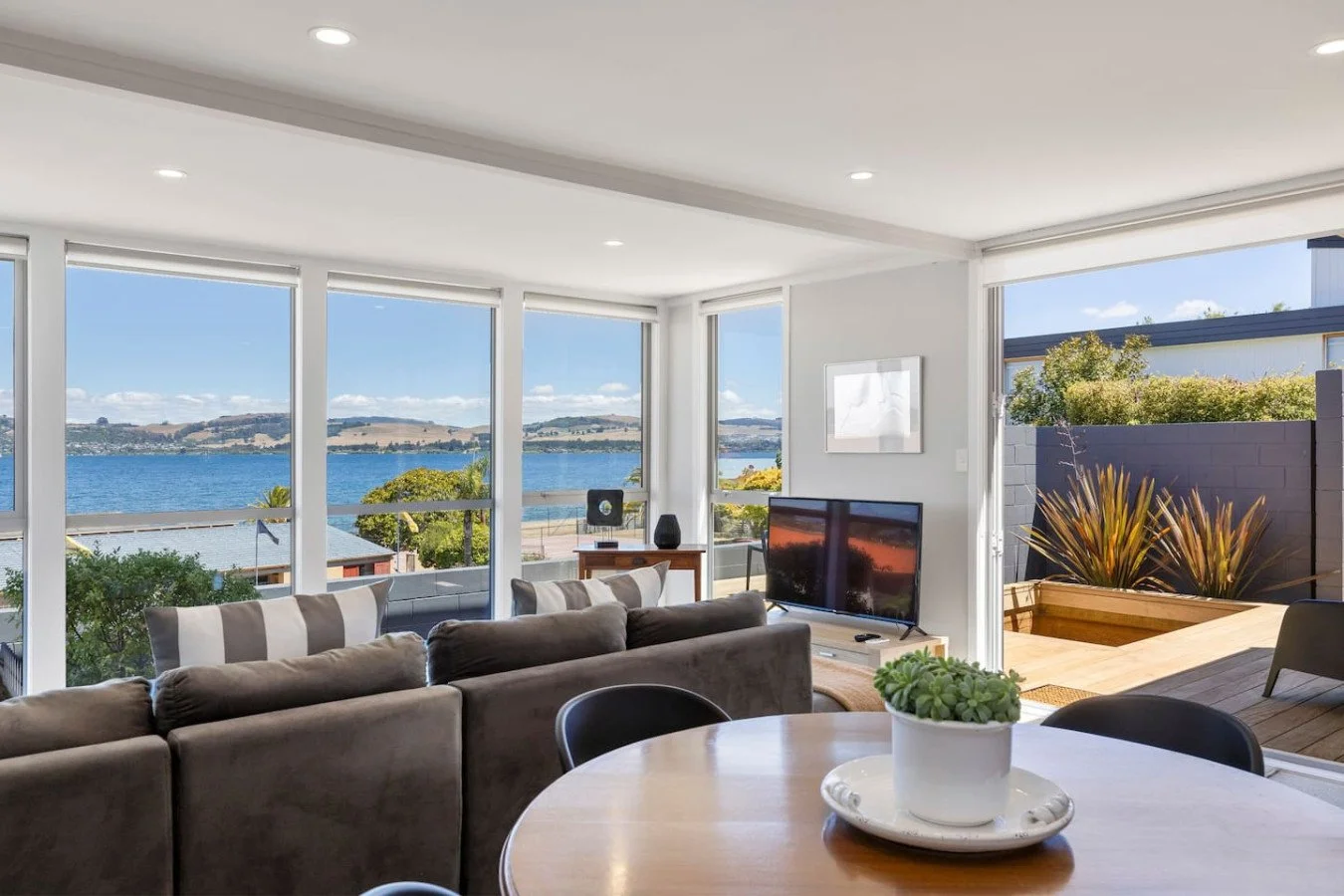 Living room with large windows overlooking a body of water and distant hills, featuring a gray sofa, a round dining table with a potted plant, a TV, and a small patio with plants outside.