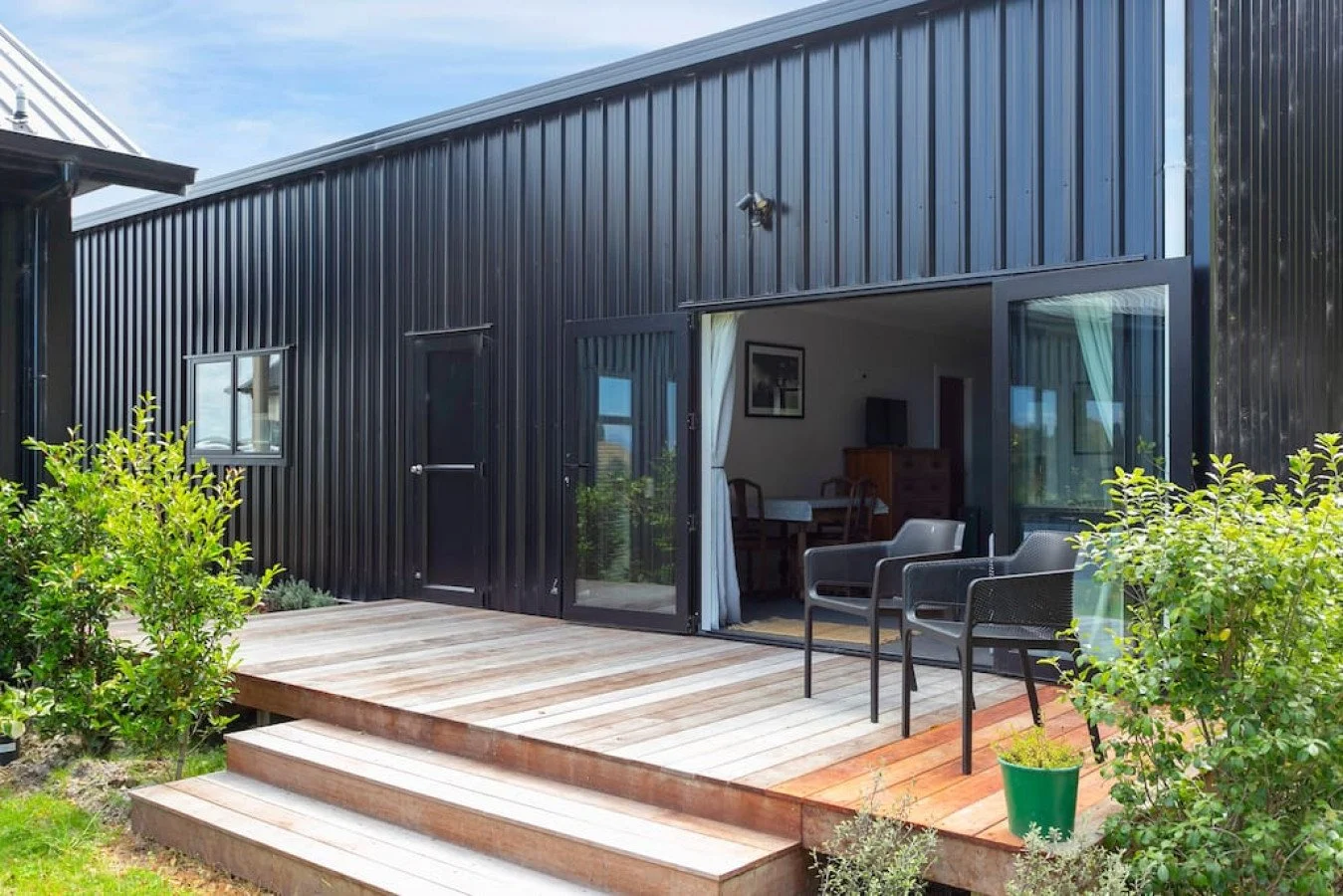 Black metal house with wooden deck, green shrubs, and outdoor seating.