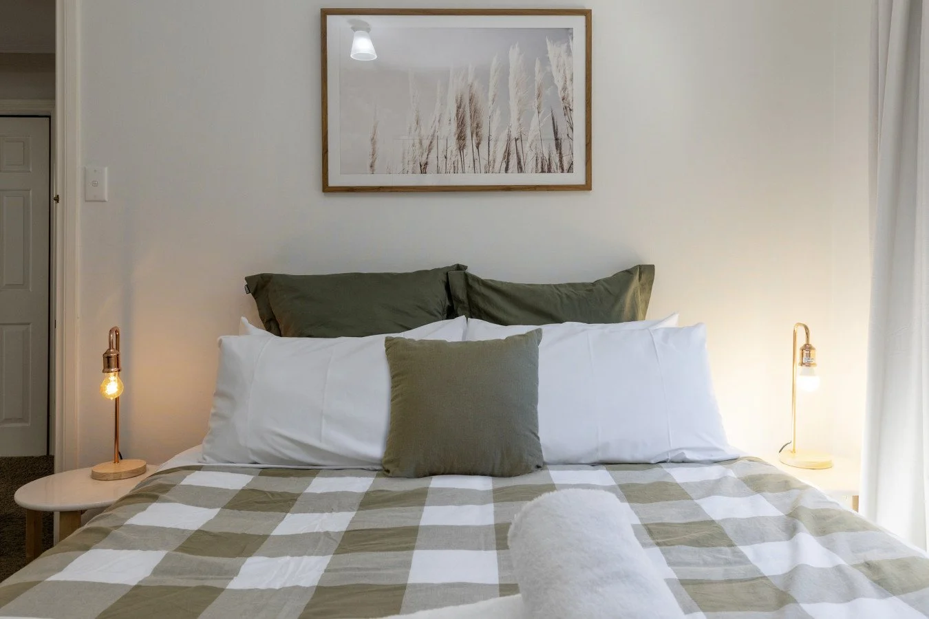 Bedroom with a neatly made bed featuring white and green pillows, a checkered bedspread, bedside lamps, and wall art above the bed.