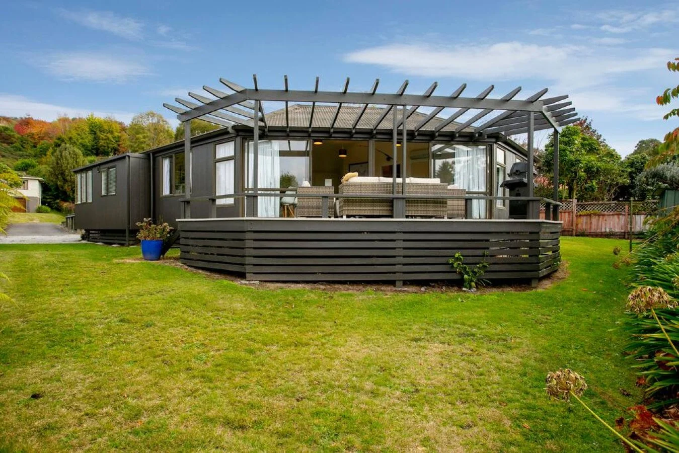 A dark gray house with a wraparound deck featuring outdoor furniture and a grill, situated on a grassy yard with trees and a hillside in the background.