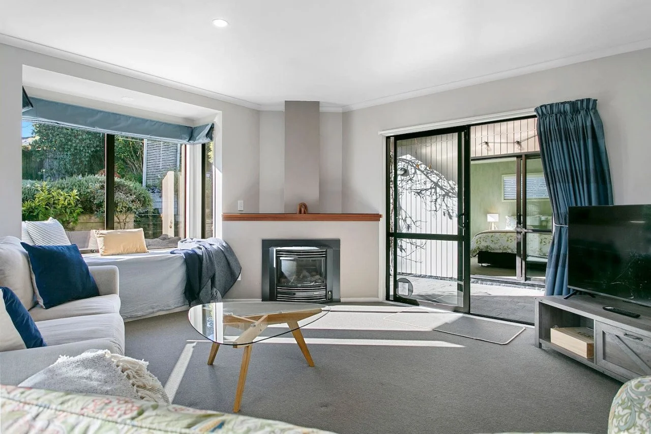 Living room with a gray sofa, blue and beige throw pillows, a glass coffee table, a fireplace, a sliding glass door with blue curtains, and a window with outdoor views.