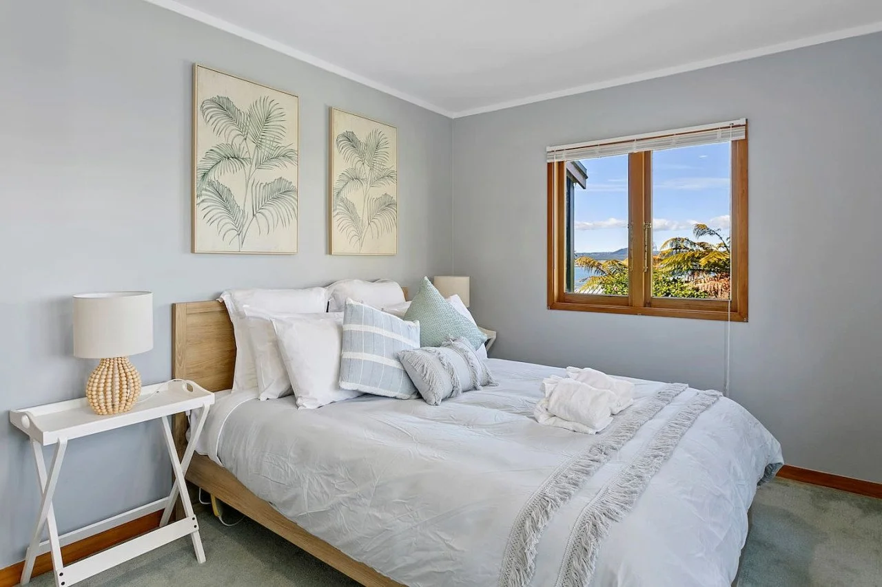 A neatly made bedroom with a wooden bed frame, white bedding, multiple pillows, two framed palm leaf art pieces on the wall, a white side table with a lamp, and a window showing a view of trees and a blue sky.