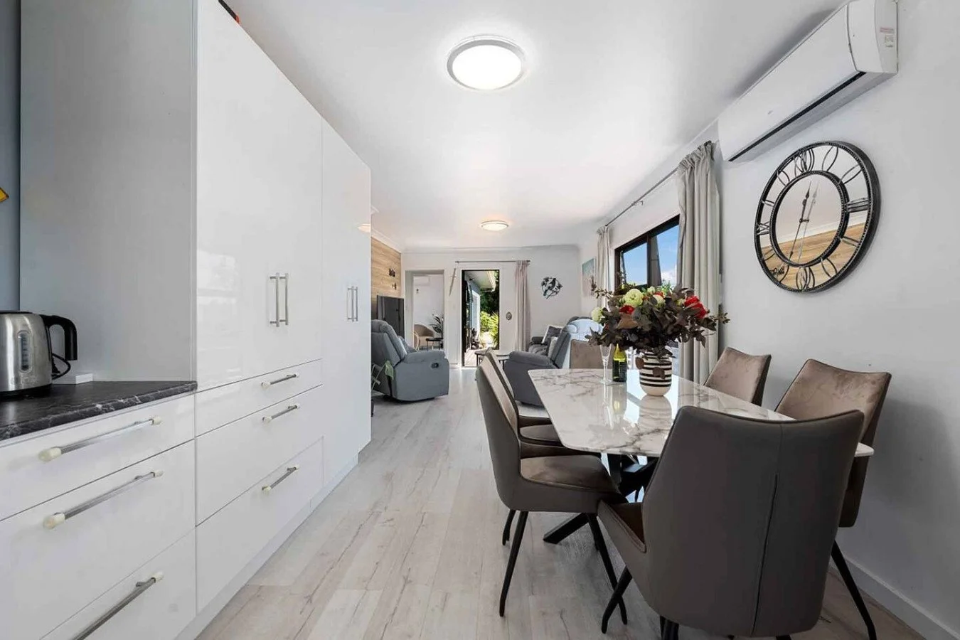 Open-concept kitchen and living room with a dining table, chairs, sofas, wall clock, window, and sliding door leading outdoors.