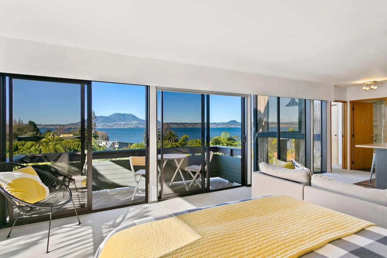 Living room with large glass sliding doors showcasing a view of a lake, mountains, and greenery. Contains a bed with a yellow quilt, a white sofa, and a balcony with outdoor furniture.
