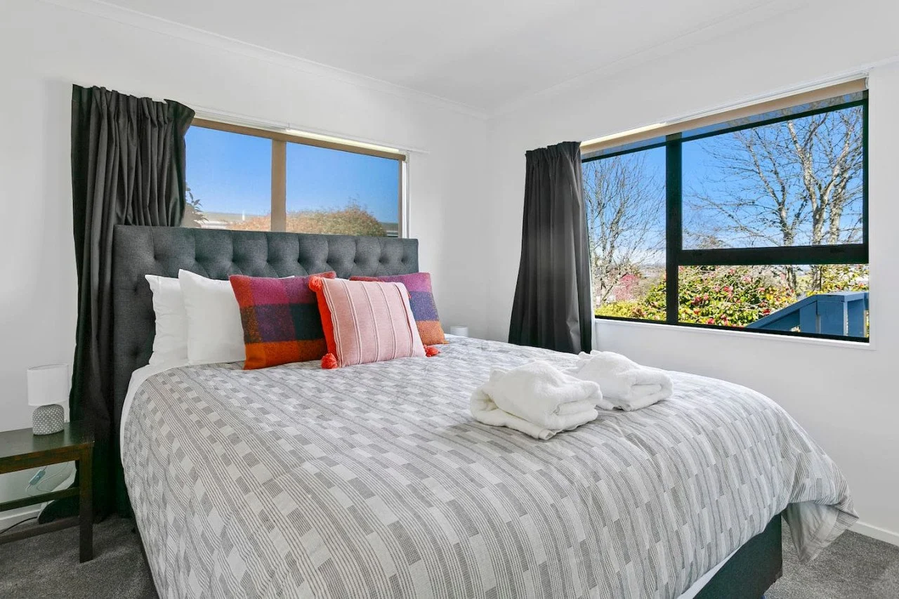 A neatly made bed in a bright bedroom with two windows showing trees and blue sky; three decorative pillows and two folded towels on the bed.