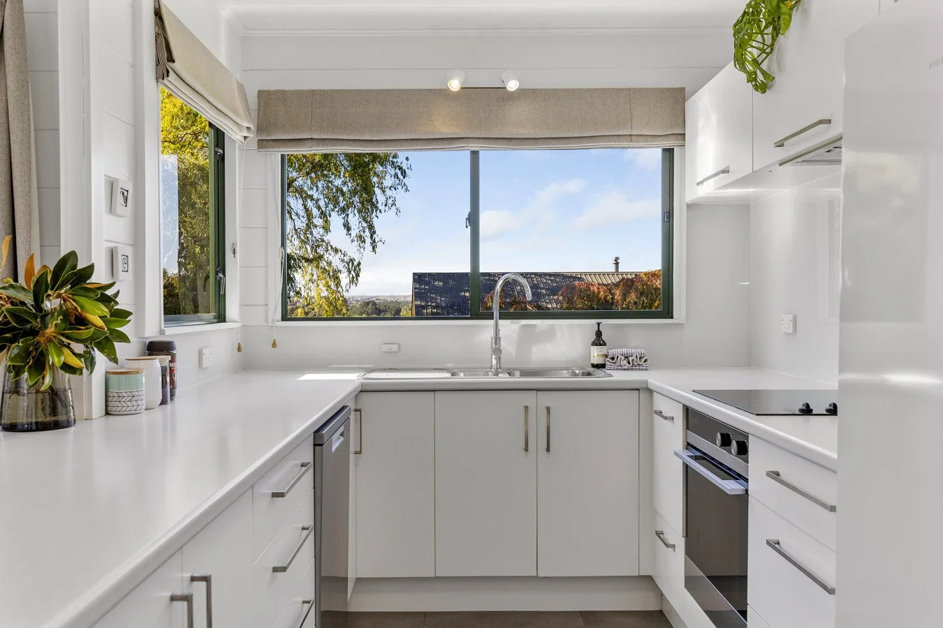 Bright white kitchen with large window showing trees and rooftops, containing white cabinetry, a sink, stove, and decorative items.