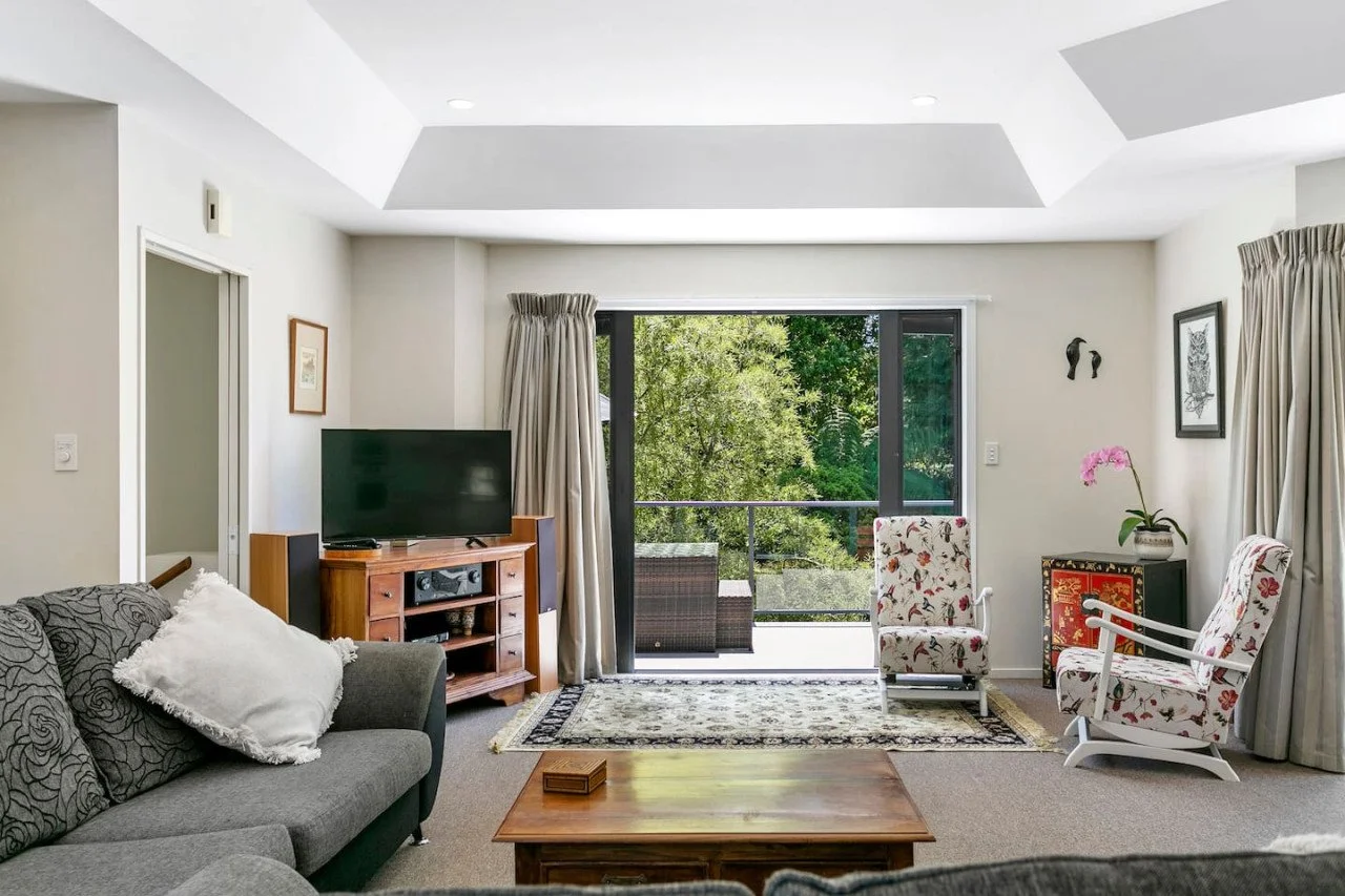 Living room with gray couch, wooden coffee table, and sliding glass door leading to balcony with outdoor seating, surrounded by trees.