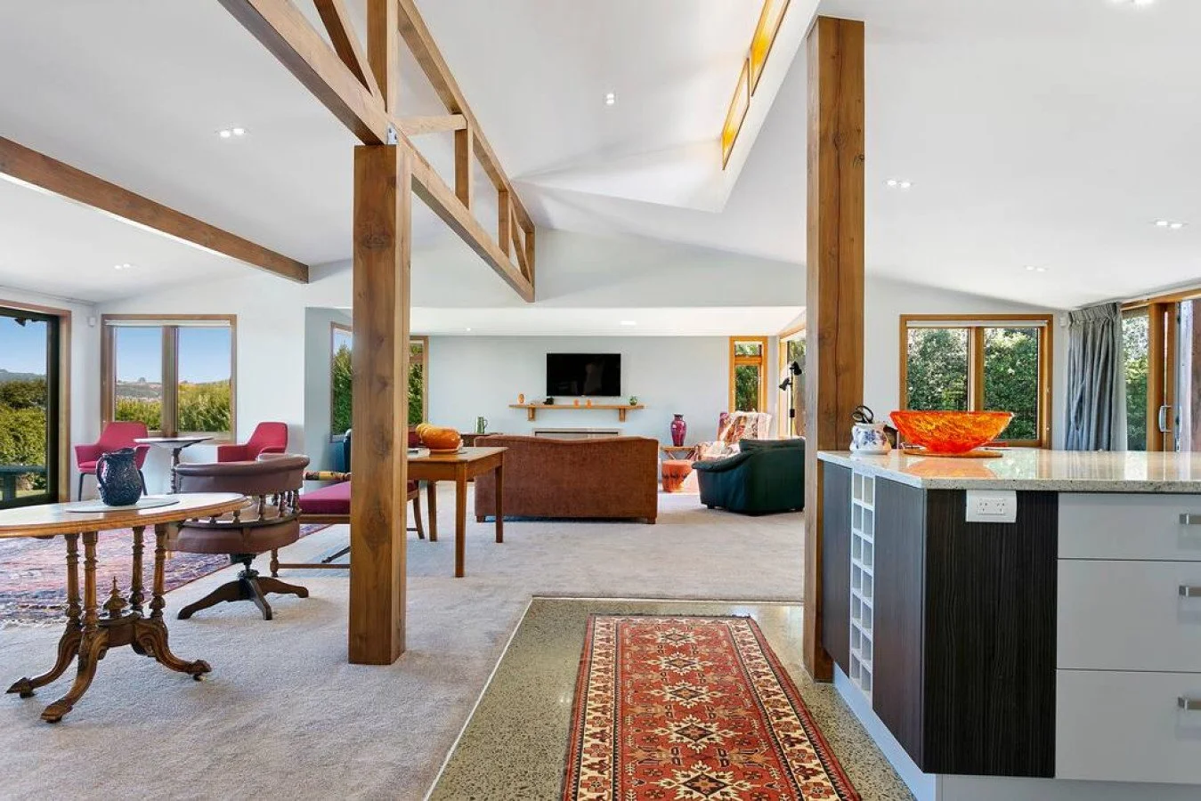 Open-concept living room and dining area with large windows, wooden beams, and various colorful furniture pieces.