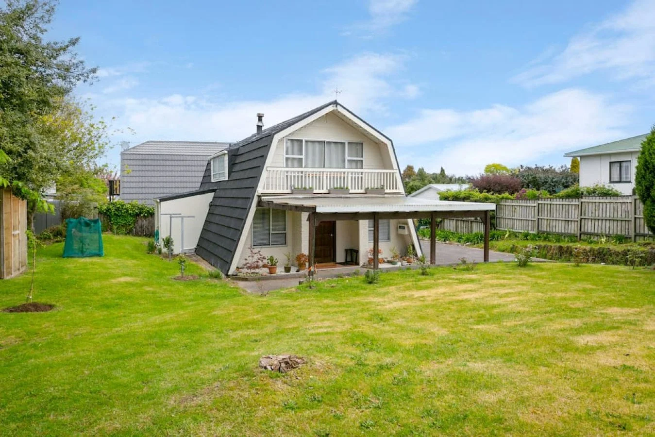 A unique two-story house with an angled roof, large front porch, and balcony overlooking a well-kept backyard with a green lawn and garden. The house has white walls, dark roof, and a small outdoor seating area.