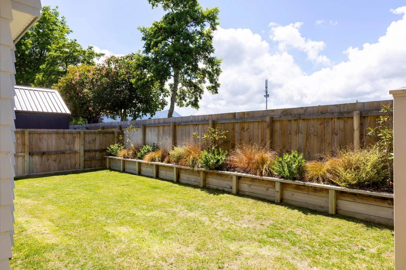 A backyard with a grass lawn, wooden fence, and a flower bed with various plants. There are trees and a cloudy sky in the background.