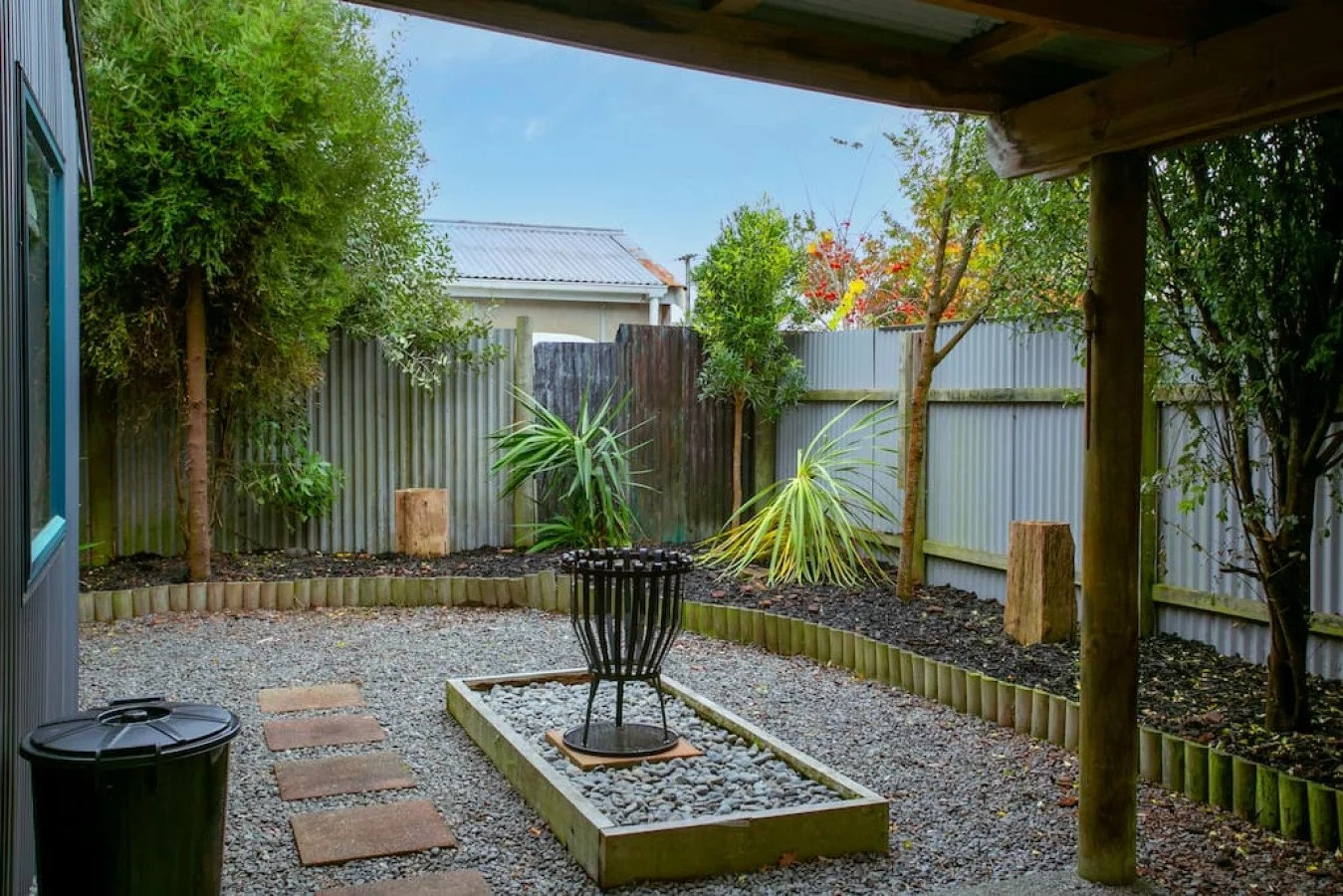 Small backyard with gravel ground, a black metal chair, a trash bin, stepping stones, and a raised garden bed with rocks. Surrounded by a metal fence, there are trees and plants, including a tall conifer and palms, with a partly cloudy sky overhead.