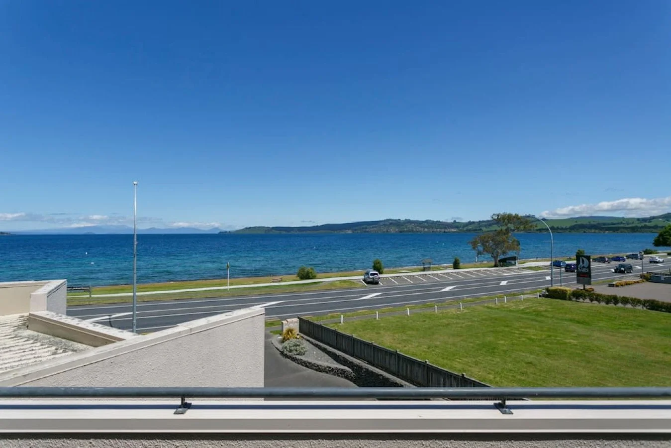 View of a coastline with a body of water, a road running parallel to the water, parked cars, a grassy area, and a clear blue sky.
