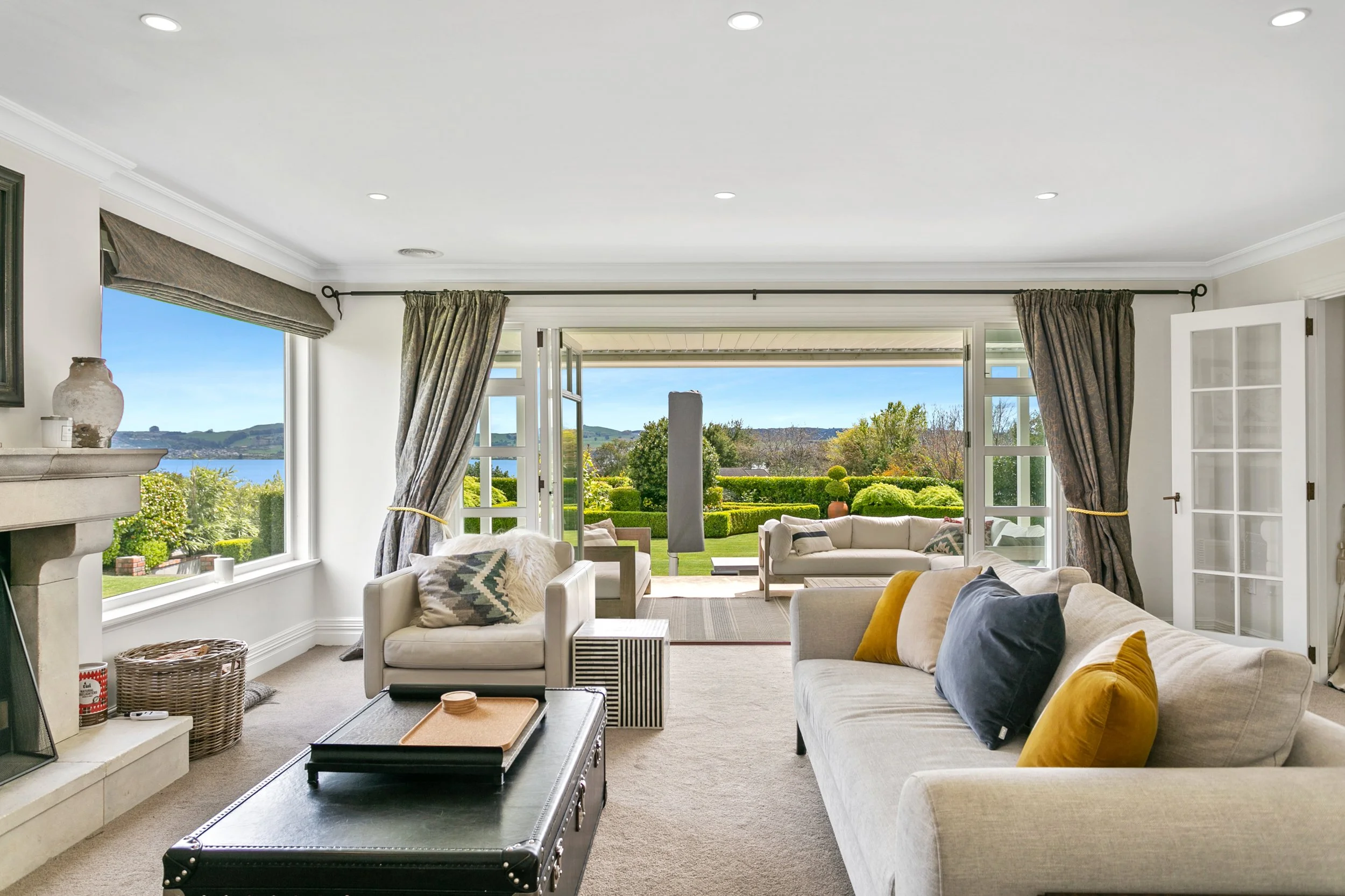 Living room with cream-colored sofas and armchairs, a black coffee table, and large windows opening to a lush garden and lake view, with distant hills.