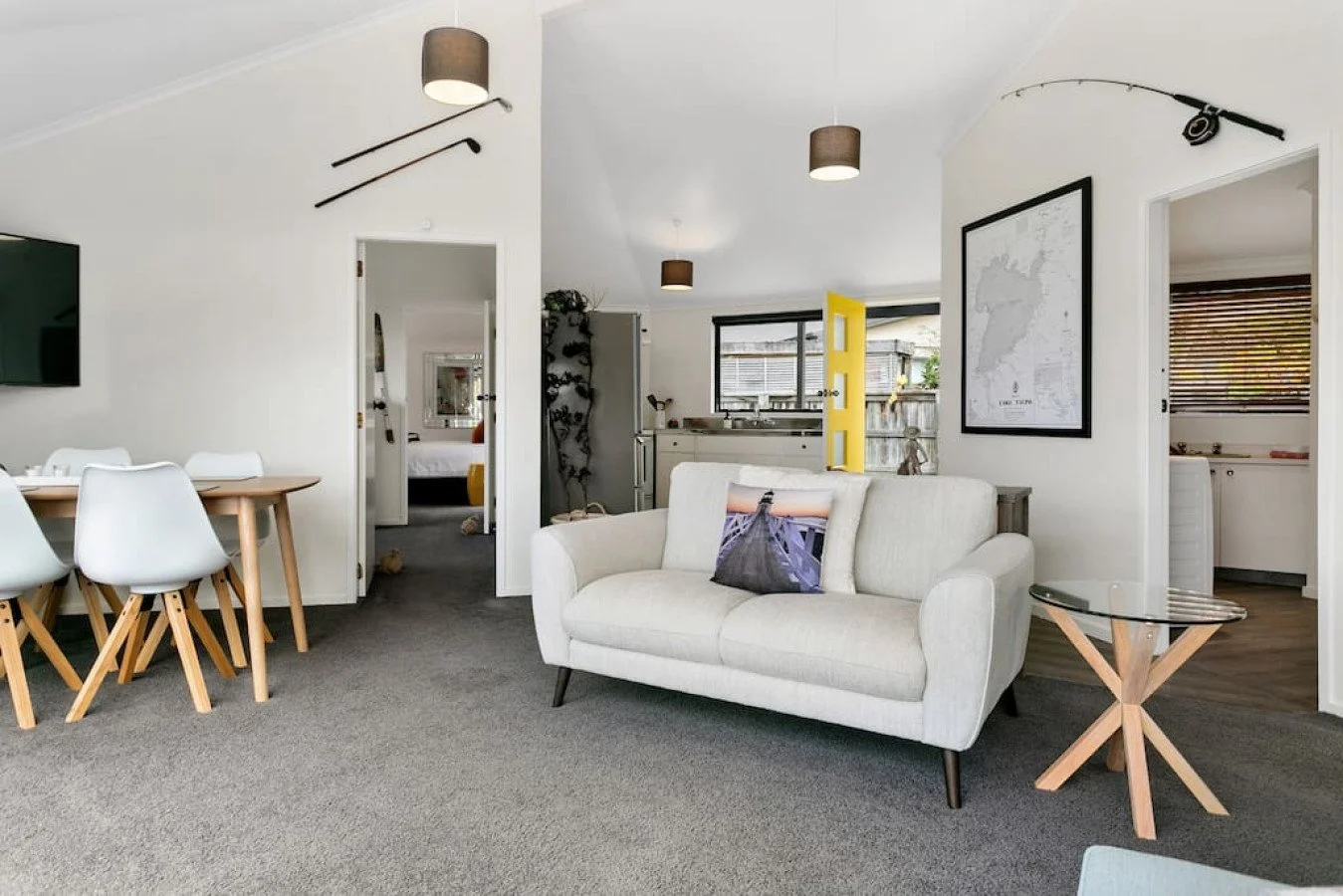 Living room with white walls, gray carpet, a light-colored sofa with a pillow, a wooden side table, and a dining area with white chairs and a wooden table. Openings reveal a kitchen, bedroom, and laundry area.