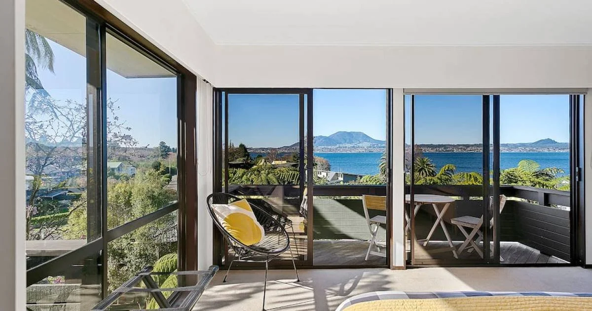 Living room with sliding glass doors opening to a balcony with ocean view, mountains, and palm trees, with outdoor chairs and a table.