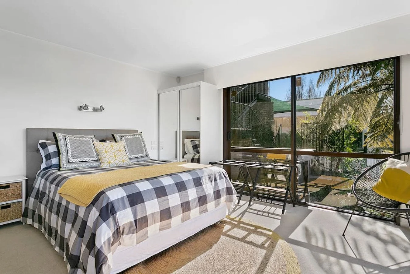 Modern bedroom with large glass sliding door overlooking a garden, bed with plaid bedding and decorative pillows, side table, chair with yellow pillow, and small outdoor table with stools.
