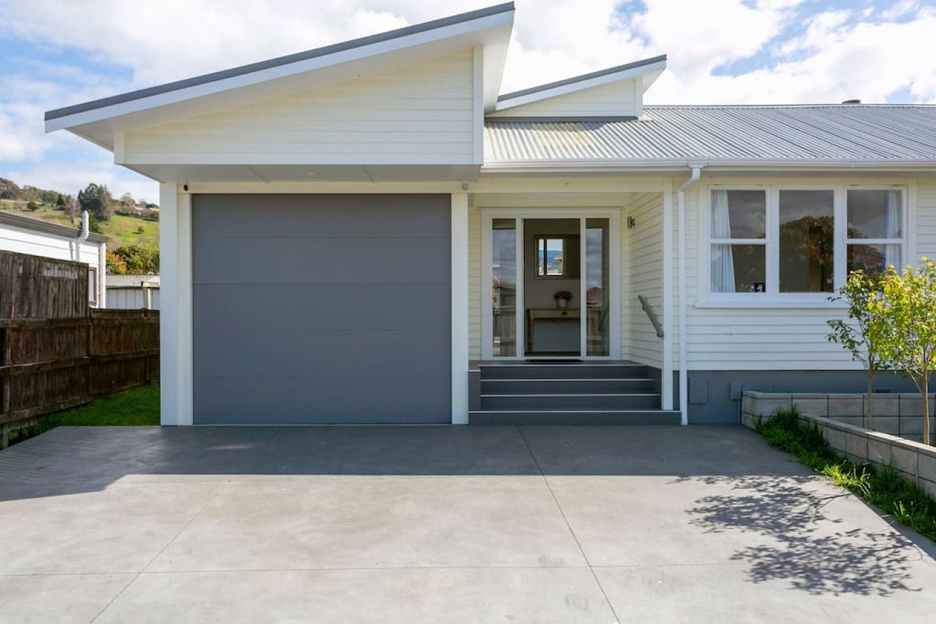 Modern suburban house with a white exterior, gray garage door, and front steps leading to a glass door, with a small garden and tree on the right side, and a concrete driveway in front.