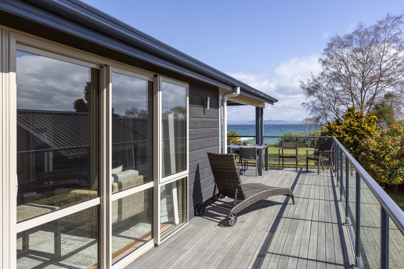 View of a house balcony with outdoor furniture, overlooking the ocean with trees and bushes in the background.