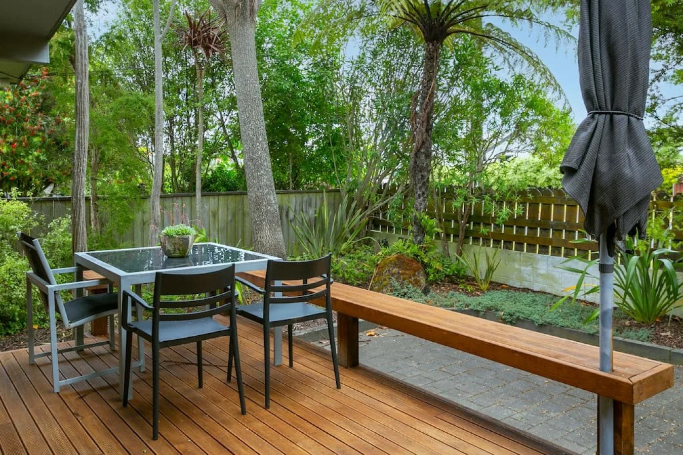 Backyard patio with wooden deck, black metal chairs around a glass-top table, a long wooden bench, a closed black patio umbrella, greenery including trees, bushes, and plants, and a wooden fence in the background.