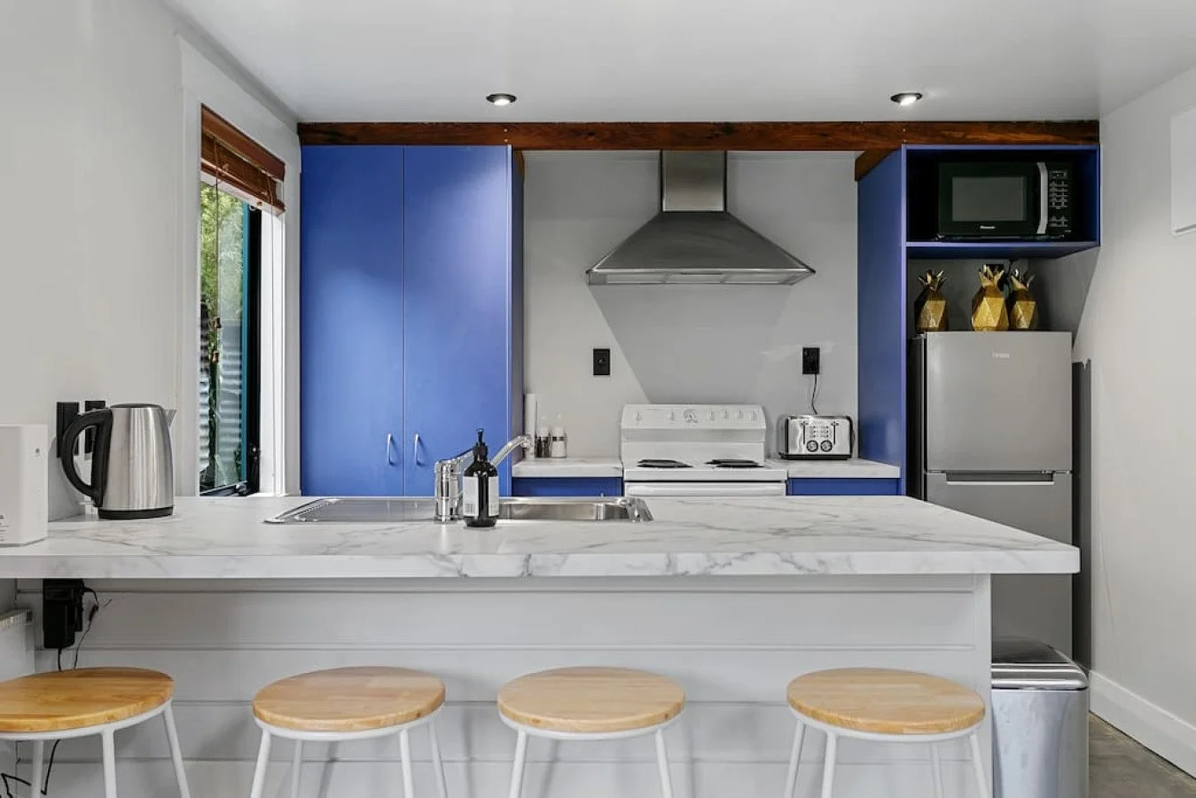 Modern kitchen with blue cabinets, stainless steel appliances, white marble countertop, and a breakfast bar with four wooden stools.