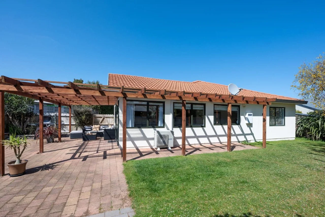 Backyard of a house with a wooden pergola, patio furniture, and a grassy lawn under a clear blue sky.