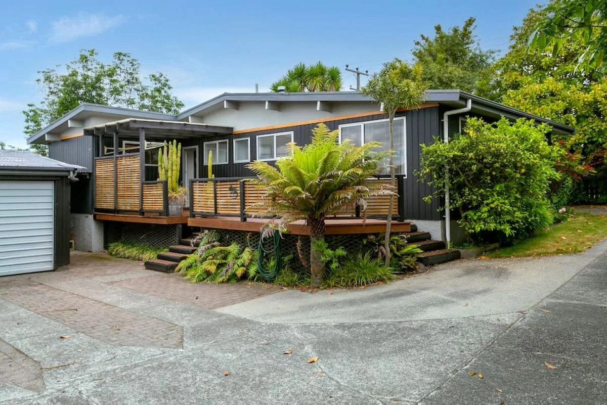 A house with a black exterior, a wooden deck with plants, steps leading up to the deck, and surrounding greenery including palm trees and bushes.