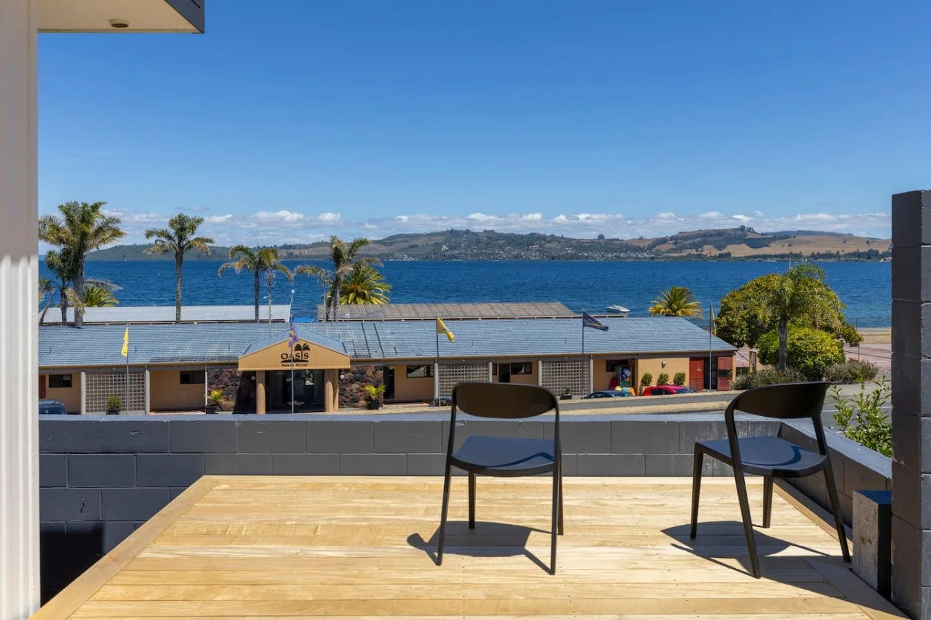 Balcony with two black chairs overlooking a lake, with trees and a building in the foreground and hills in the background.