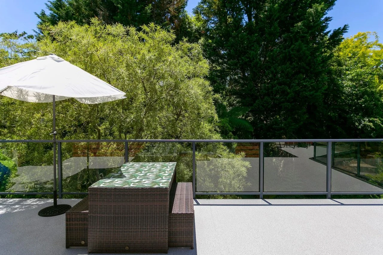 Balcony with wicker furniture, a patterned cushion, a white umbrella, and green trees in the background under a clear blue sky.