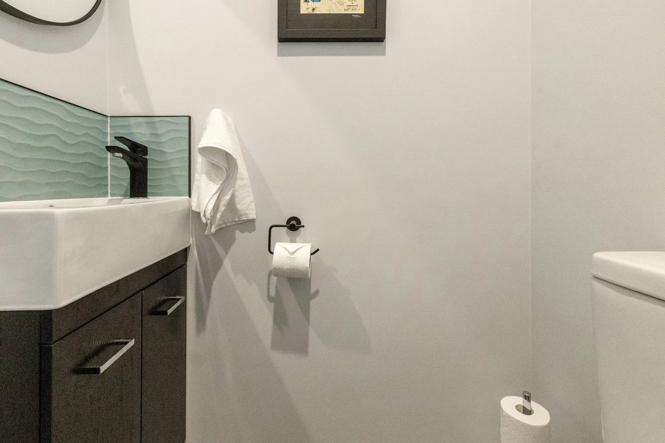A modern bathroom with a white wall, a black sink faucet, a black vanity, a white sink, a towel hanging on a wall hook, a toilet paper roll, and a toilet with a closed lid.