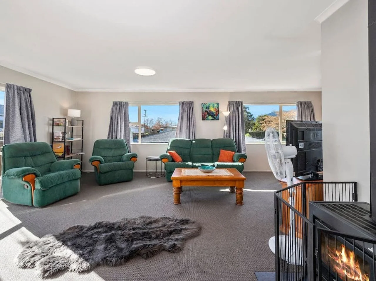 Living room with green upholstered chairs, a wooden coffee table, a gray rug, a fireplace, and large windows with gray curtains offering a view of the outdoors.