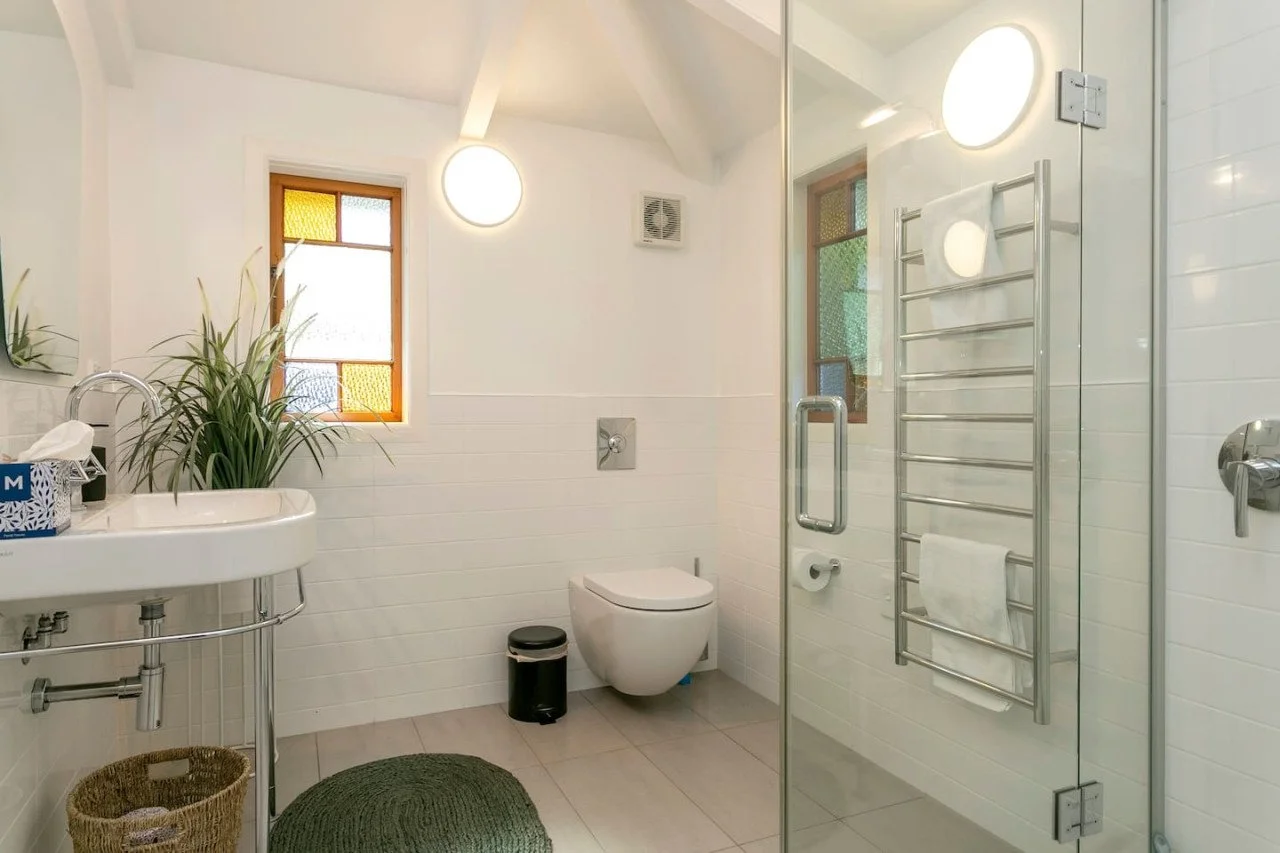 Clean, modern bathroom with white walls, a small window, and a glass shower enclosure. Includes a white sink with a plant, a toilet, a small trash can, and a heated towel rack.