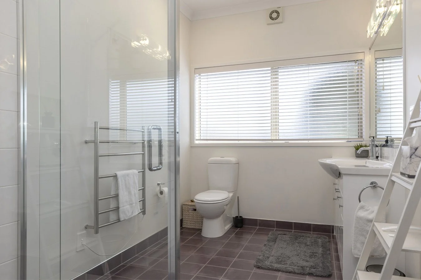 A clean bathroom with a glass shower enclosure, a toilet, a sink, and a large window with blinds. There are gray tiles on the floor and white walls.