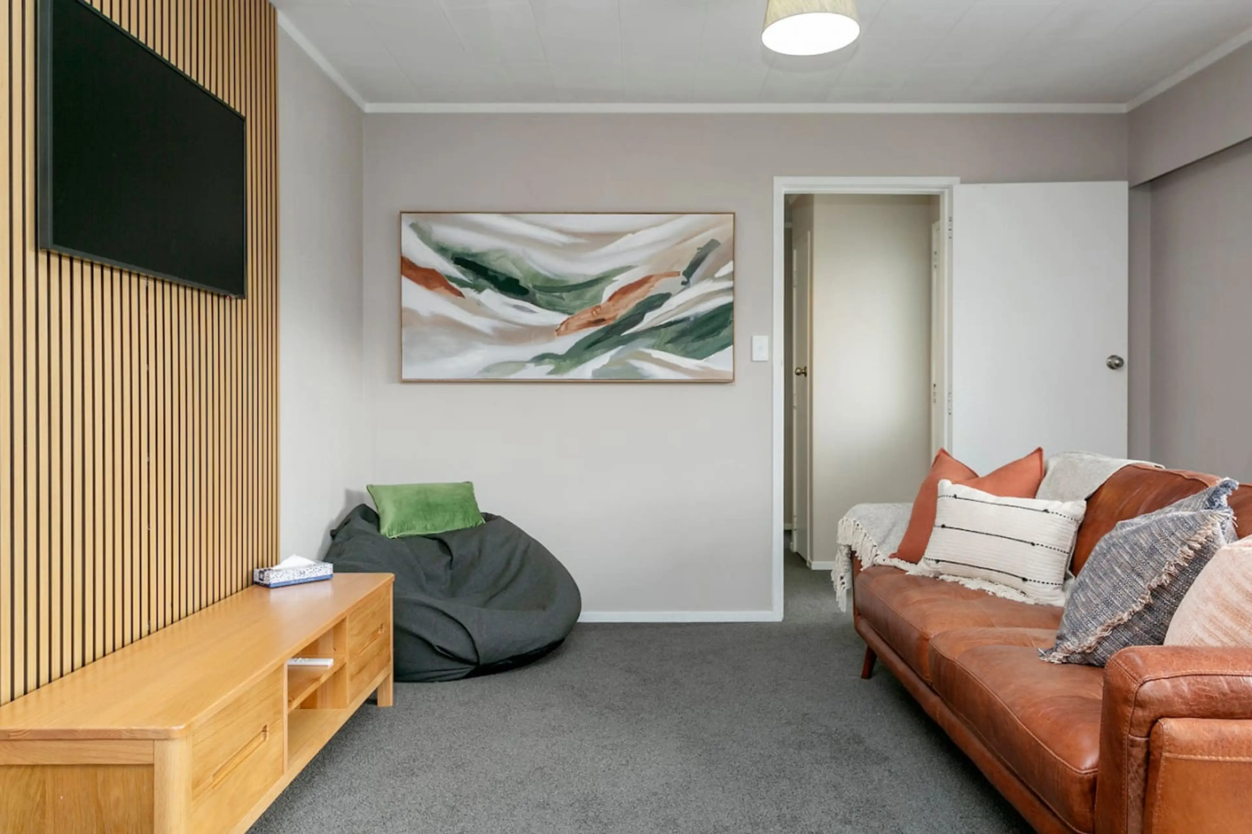 Living room with a leather sofa with pillows, a wooden TV stand, a large wall-mounted TV, and an abstract wall art. There's a gray bean bag with a green cushion and a door leading to another room.