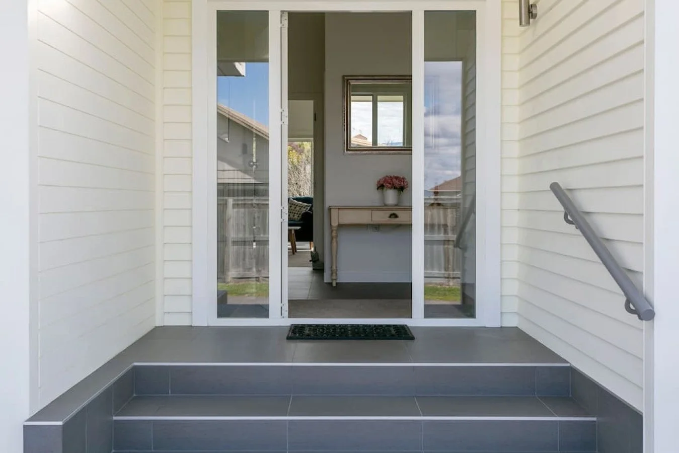 Front porch with sliding glass door leading inside a house, with a black doormat on gray tiled steps and a gray railing on the side.