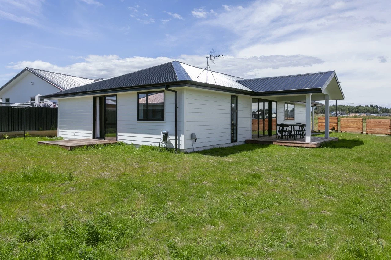 A modern single-story house with a white exterior and black roof, featuring a small front porch with outdoor furniture, surrounded by a green lawn and a wooden fence under a partly cloudy sky.
