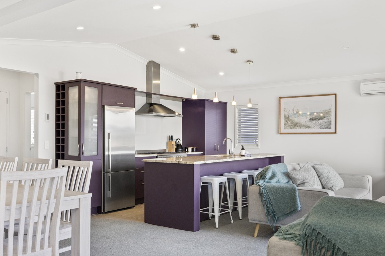 Open-concept living space with kitchen and dining area, featuring purple cabinetry, stainless steel refrigerator, kitchen island with bar stools, and a adjacent seating area with a white couch and wall art.