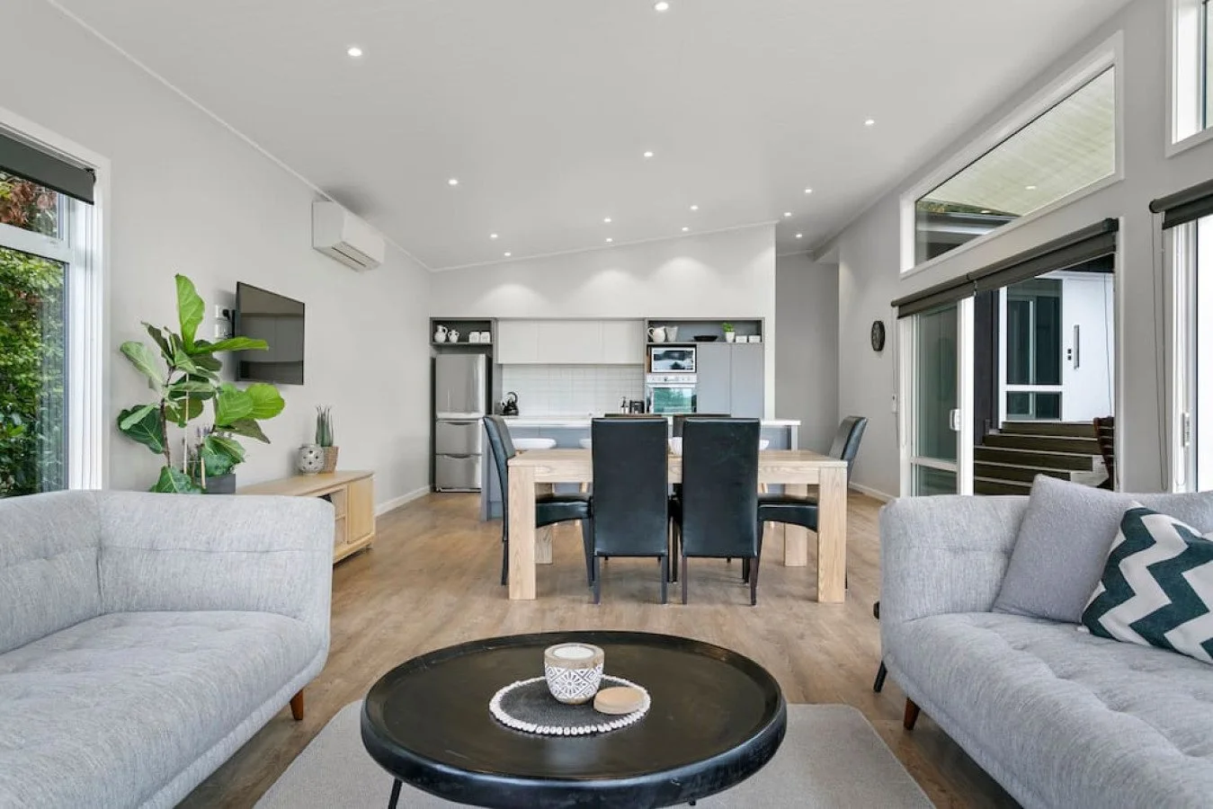 Open-concept living and dining area with modern furnishings, white walls, and large windows letting in natural light. The living room has two light gray sofas, a black coffee table, and a plant. The dining area features a wooden table with six black 