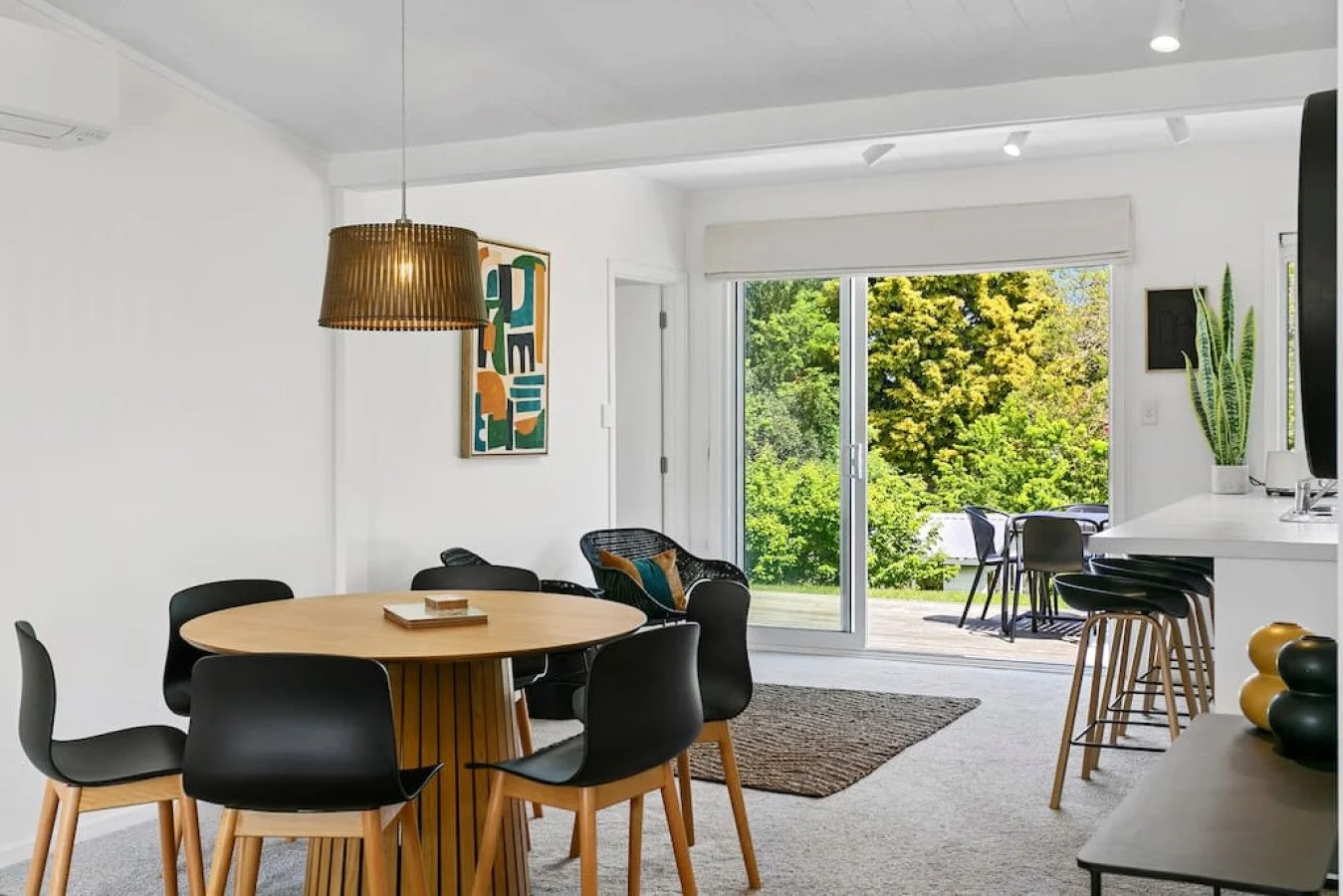 Open-concept living and dining area with sliding glass door leading to outdoor deck surrounded by green trees. Dining table with six black chairs, bar area with stools, and a potted plant inside.