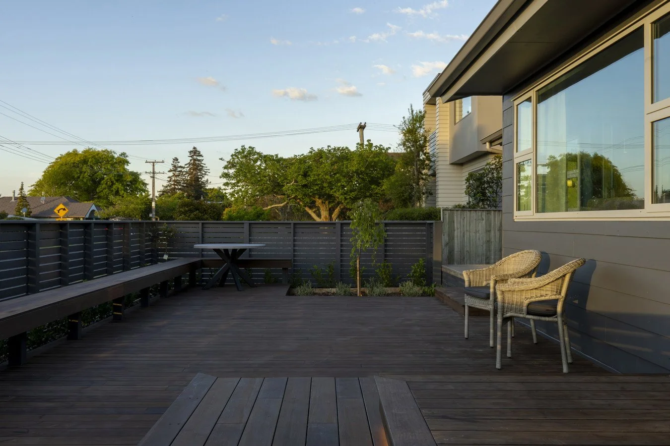 A wooden deck patio with outdoor chairs and a small table, enclosed by a dark gray fence, with trees and houses in the background under a partly cloudy sky.