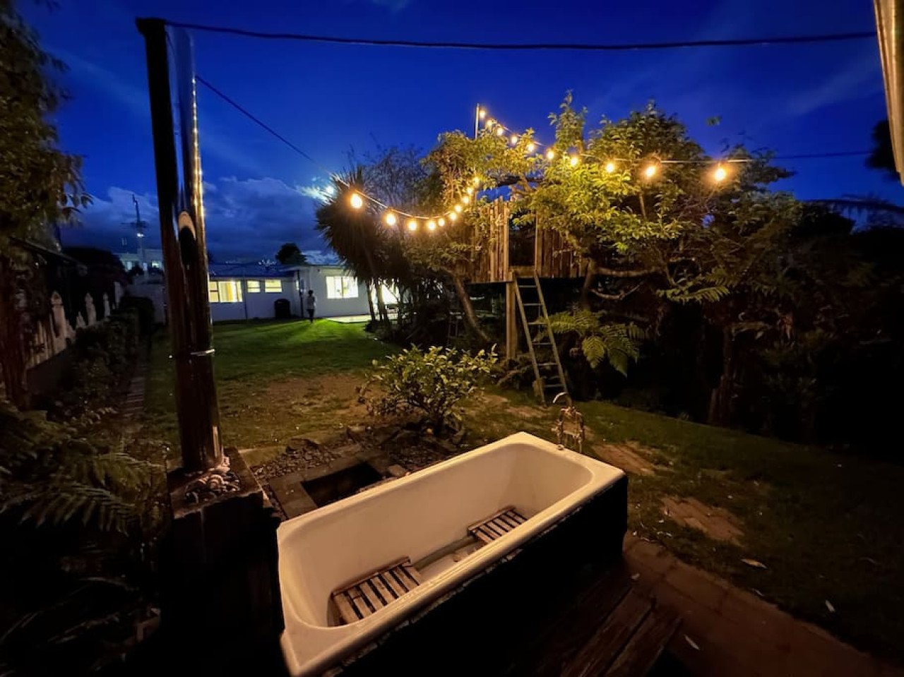 Outdoor backyard scene at dusk with string lights hanging between trees, a wooden treehouse with a ladder, a garden with bushes, and a vintage bathtub with wooden slats inside placed on a brick pathway.