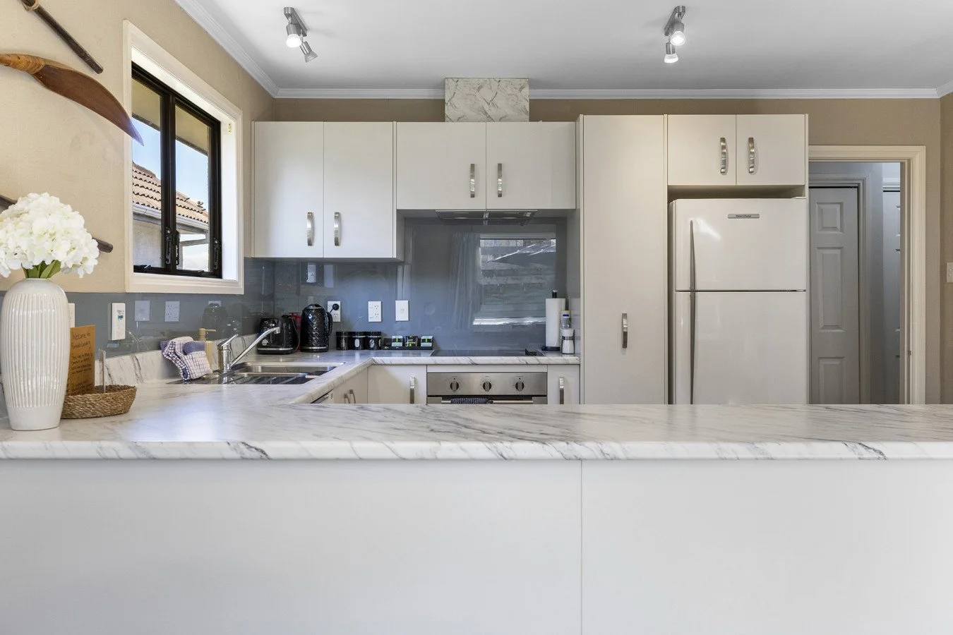 Modern kitchen with white cabinets, marble countertop, stainless steel appliances, a window with a view of neighboring rooftops, and minimal decor including a vase with flowers and small kitchen appliances on the counter.