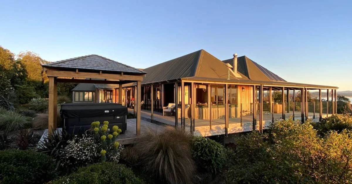 A house with wood siding and a metal roof, surrounded by a porch, with a hot tub and garden in the foreground, overlooking a sunny landscape.