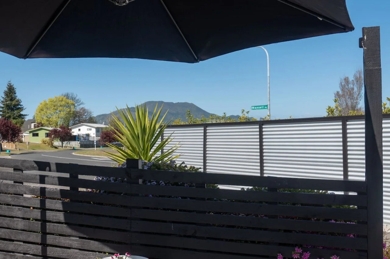 A backyard patio with a black umbrella, a black wooden fence, a large green plant, and neighboring houses with trees and mountains in the background.