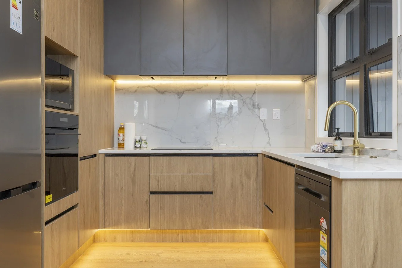 Modern kitchen with wooden cabinetry, white marble backsplash, and gray upper cabinets, featuring a gold faucet and large window.