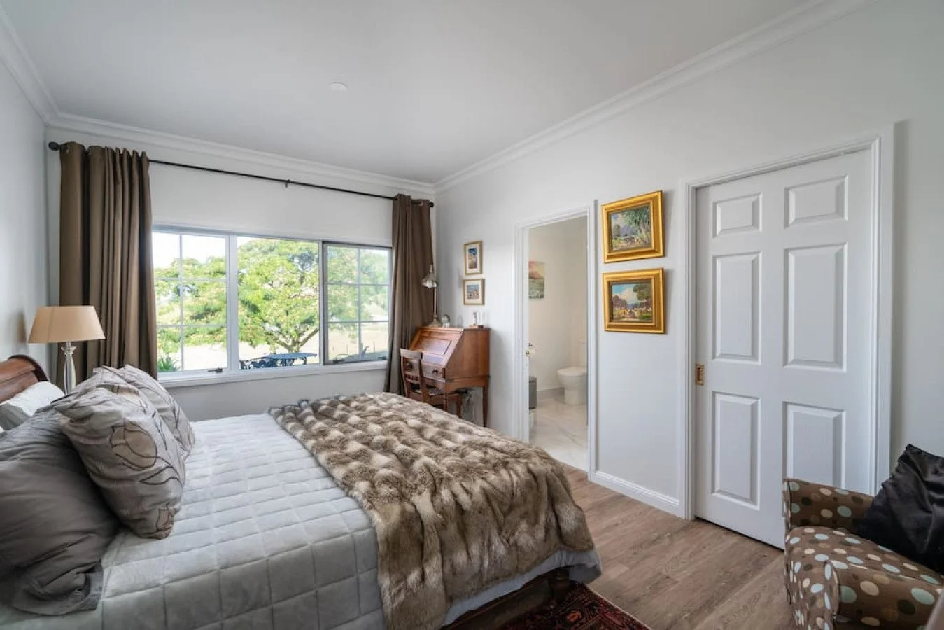 A bedroom with a large window, brown curtains, a bed with patterned pillows and a faux fur blanket, a wooden writing desk, and a small bathroom visible through the open door.