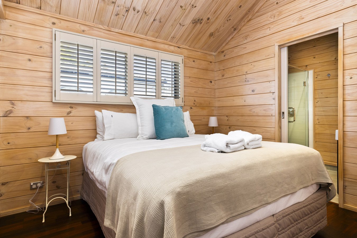 A cozy bedroom with wood-paneled walls and ceiling, a neatly made bed with white and beige linens, a blue accent pillow, and folded white towels on top, flanked by two small white nightstands with lamps, and a window with shutters above the bed.