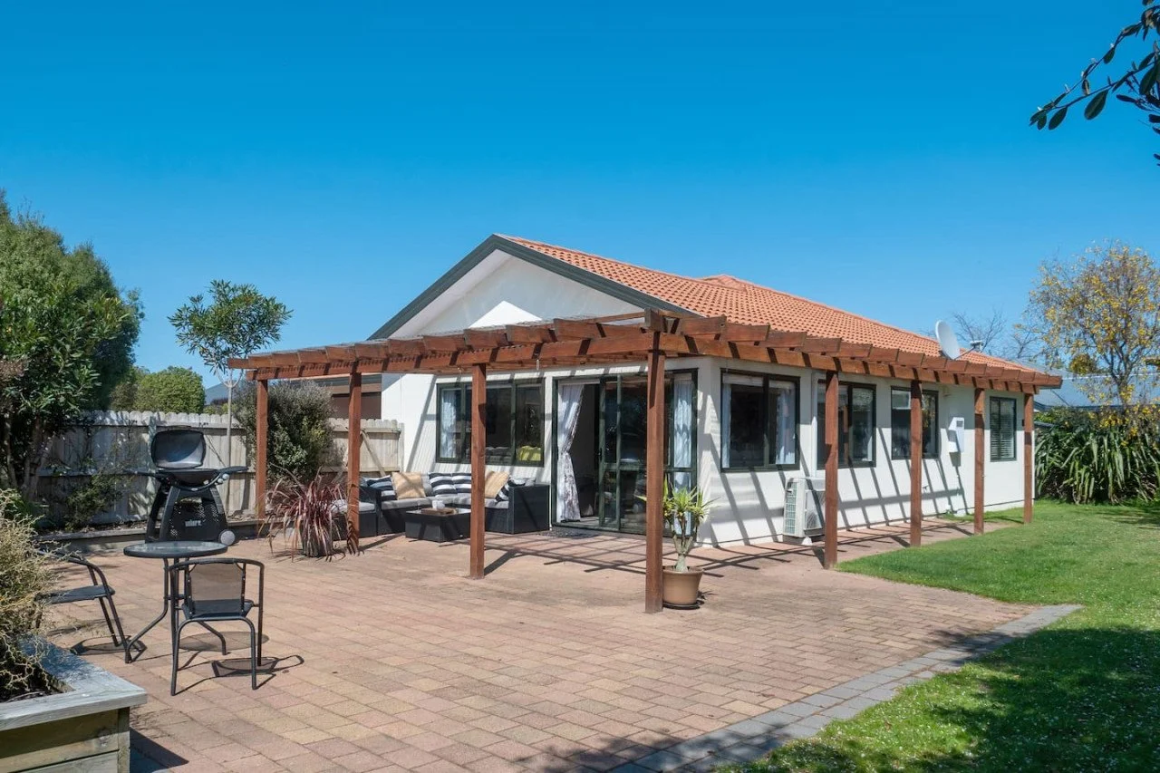 Backyard patio with wooden pergola, outdoor seating, barbecue grill, and garden, adjacent to a white house with orange roof tiles under a clear blue sky.