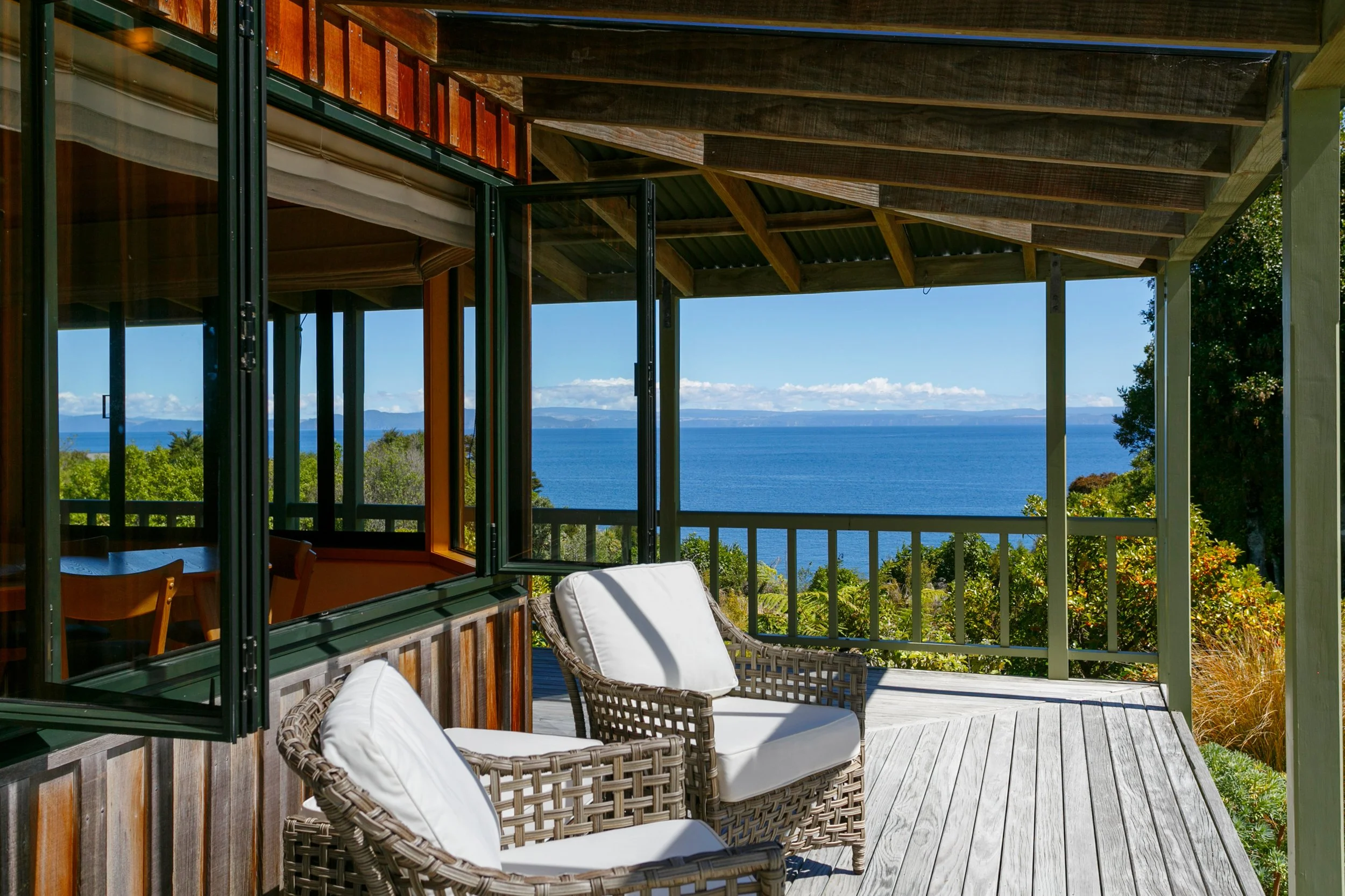 View from a porch with wicker chairs and white cushions, overlooking a body of water and lush green trees under a partly cloudy sky.