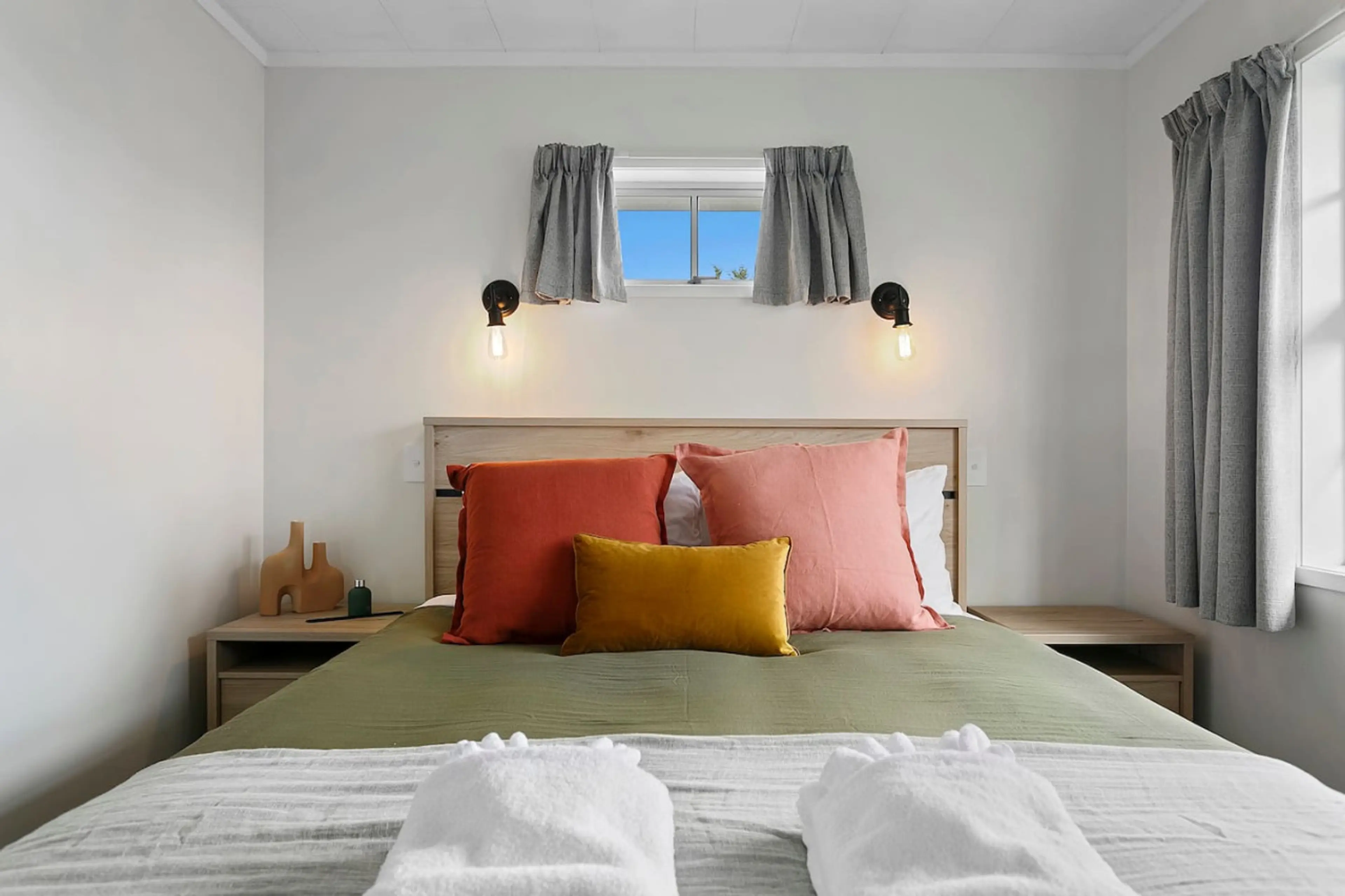 Bedroom with a bed, pink and orange pillows, green cover, wooden headboard, nightstands, decorative vases, wall-mounted lamps, window with curtains, view of blue sky outside.